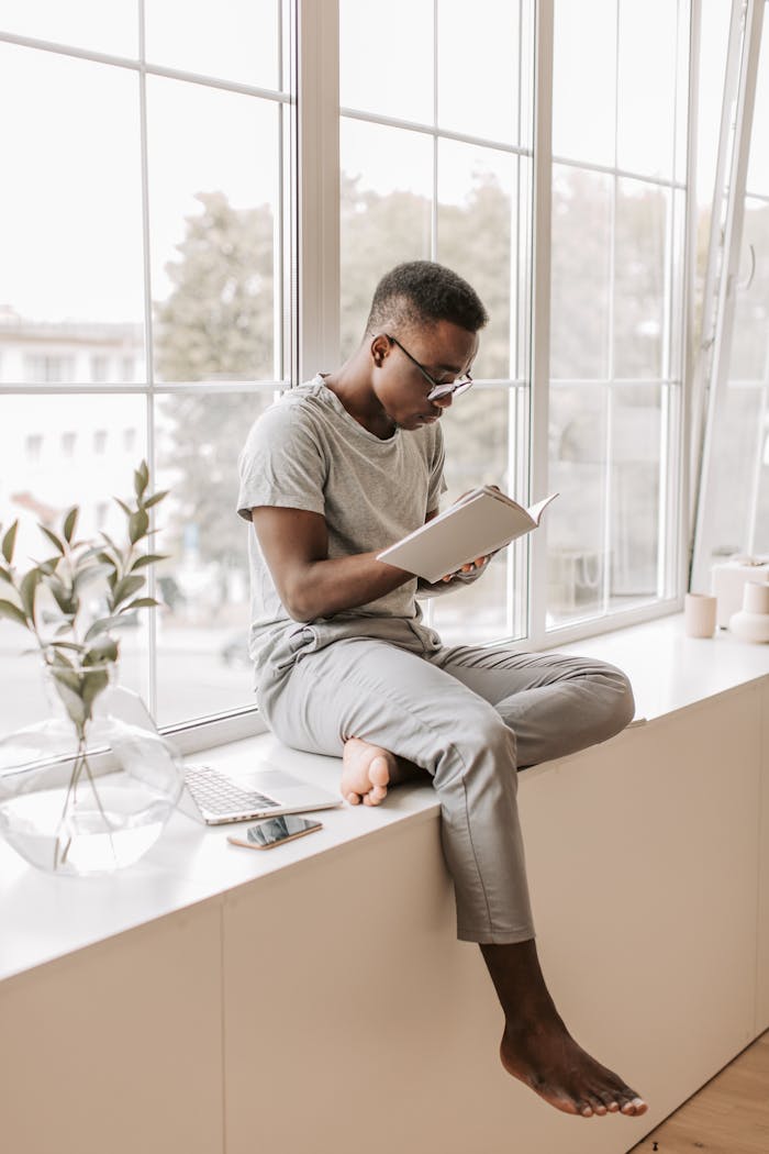 A young man enjoys a book while sitting on a sunlit window sill indoors.