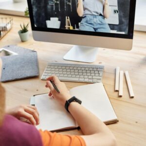 Student attending virtual class on a computer, taking notes at home.